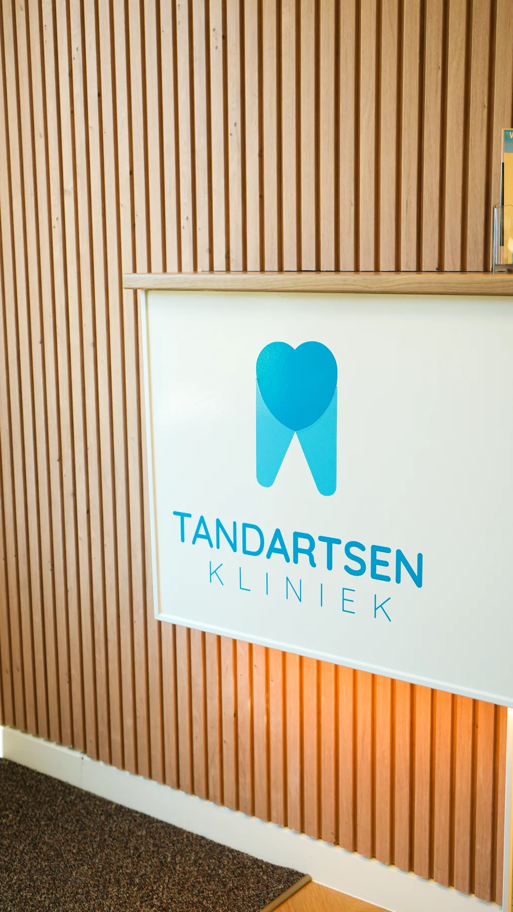 Logobord Tandartsen Kliniek op de houten lamellenwand bij de entree.