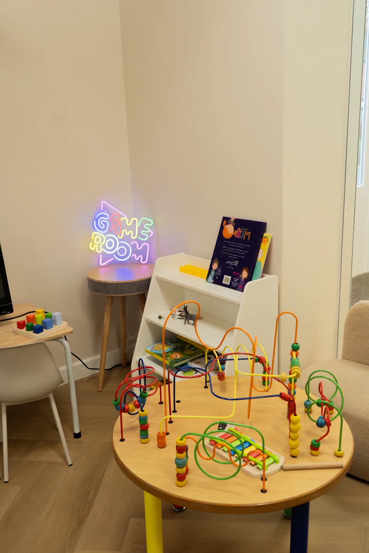 Speelhoek met kralenspel, boekenkastje en een neonbordje 'Game Room'.